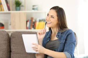 Pensive girl holding notebook looking at side sitting on a sofa in the living room at home